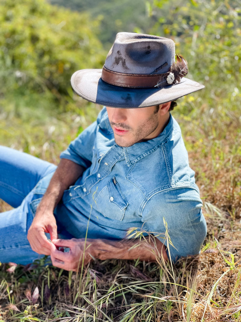 A man leaning on the grass wearing a denim jacket and a fedora hat