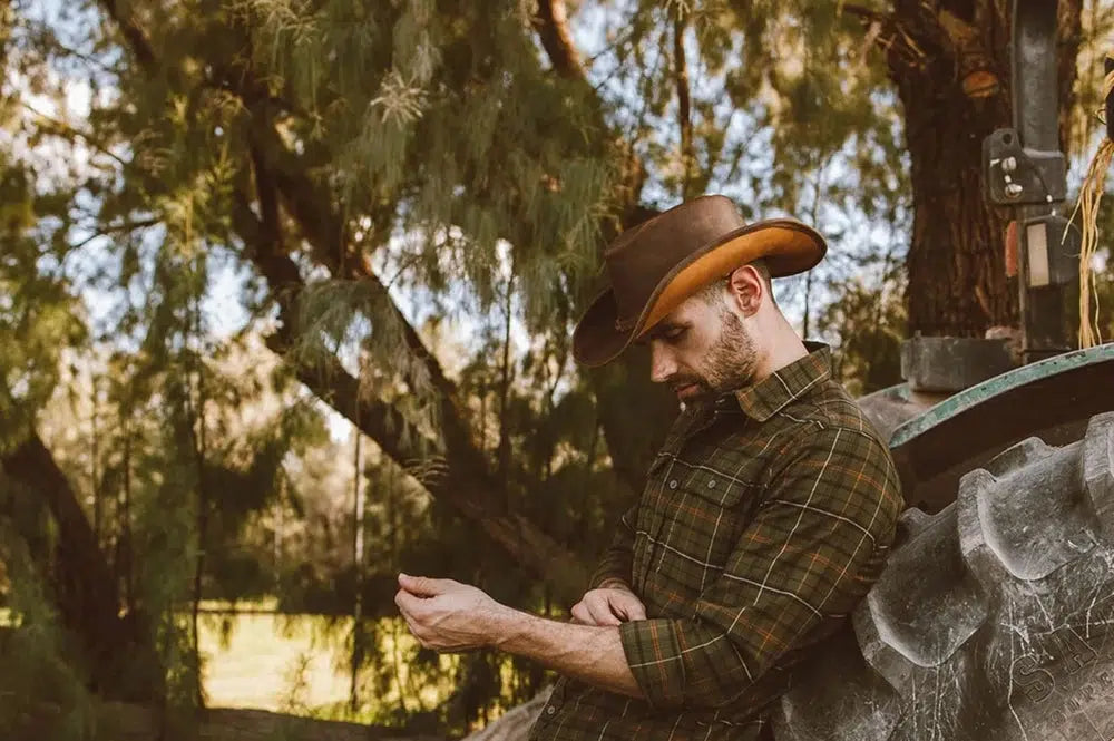 A man staying near a lake wearing a plaid shirt and brown leather hat