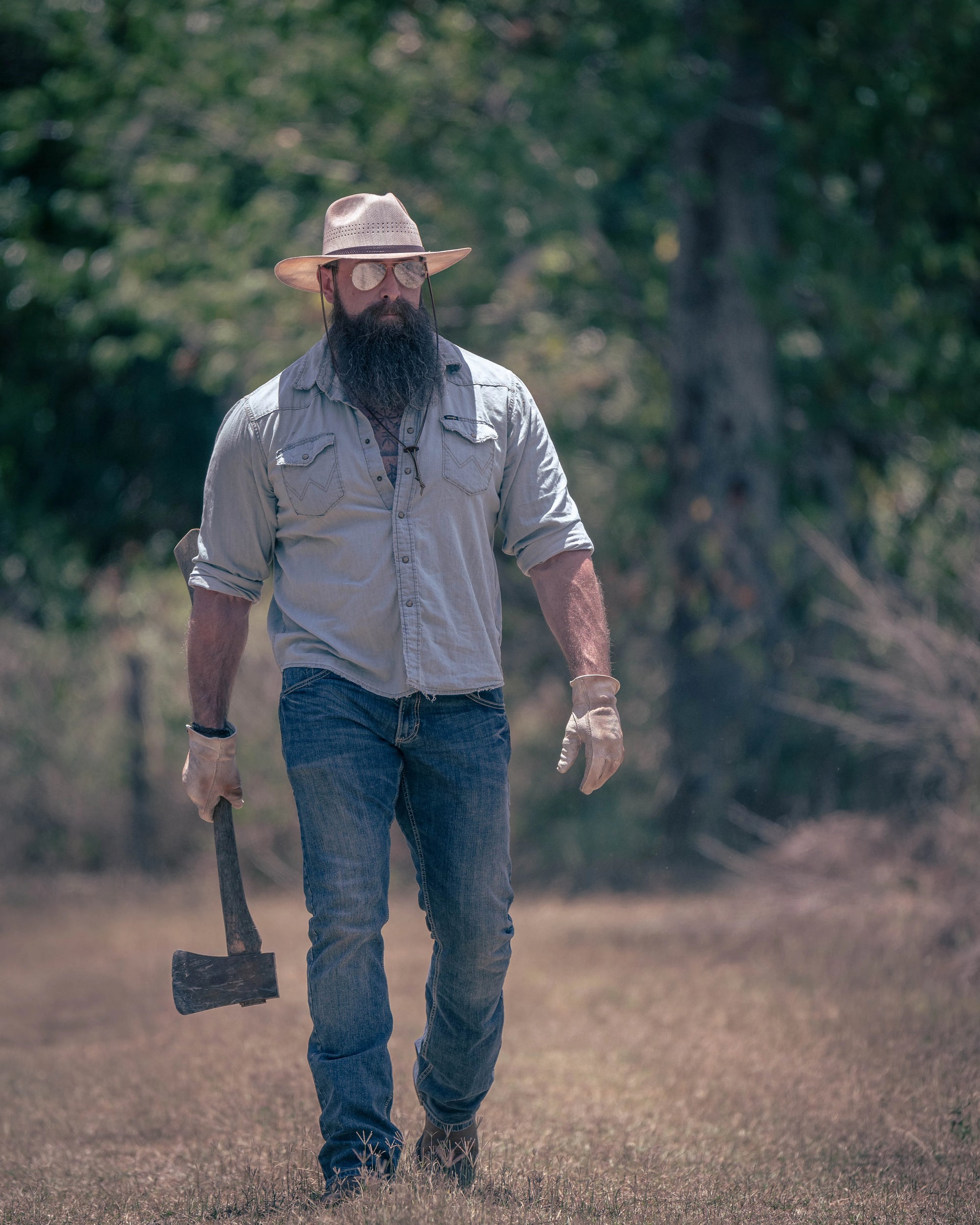 A man with a sun hat walking outdoors carrying an axe