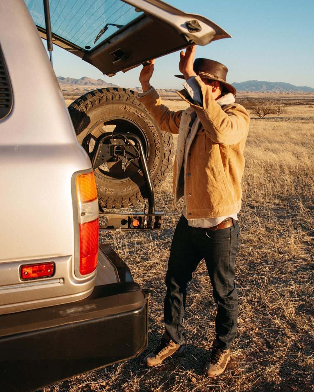 A man wearing a brown cowboy hat and jacket standing opening a car's back door