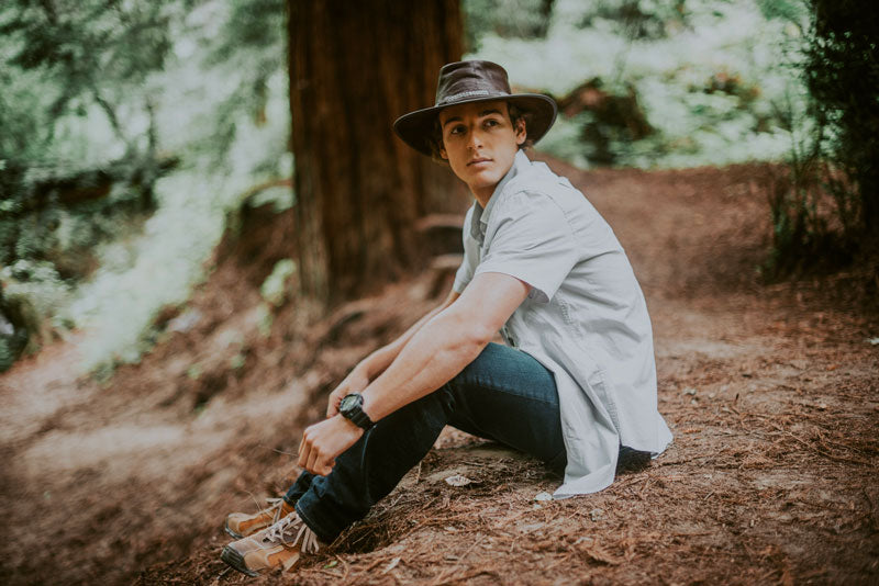 A man on the forest sitting on the ground wearing a bison leather hat