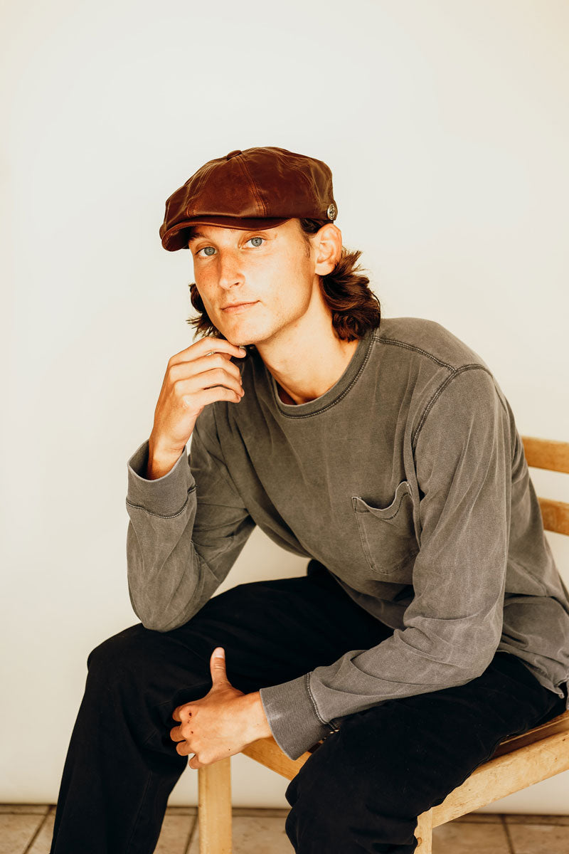 A man sitting down wearing a gray sweatshirt and a brown leather cap