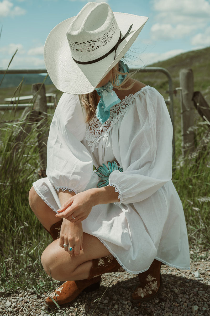 A woman squatting outdoors wearing a light blue scarf and a cream straw cowboy hat