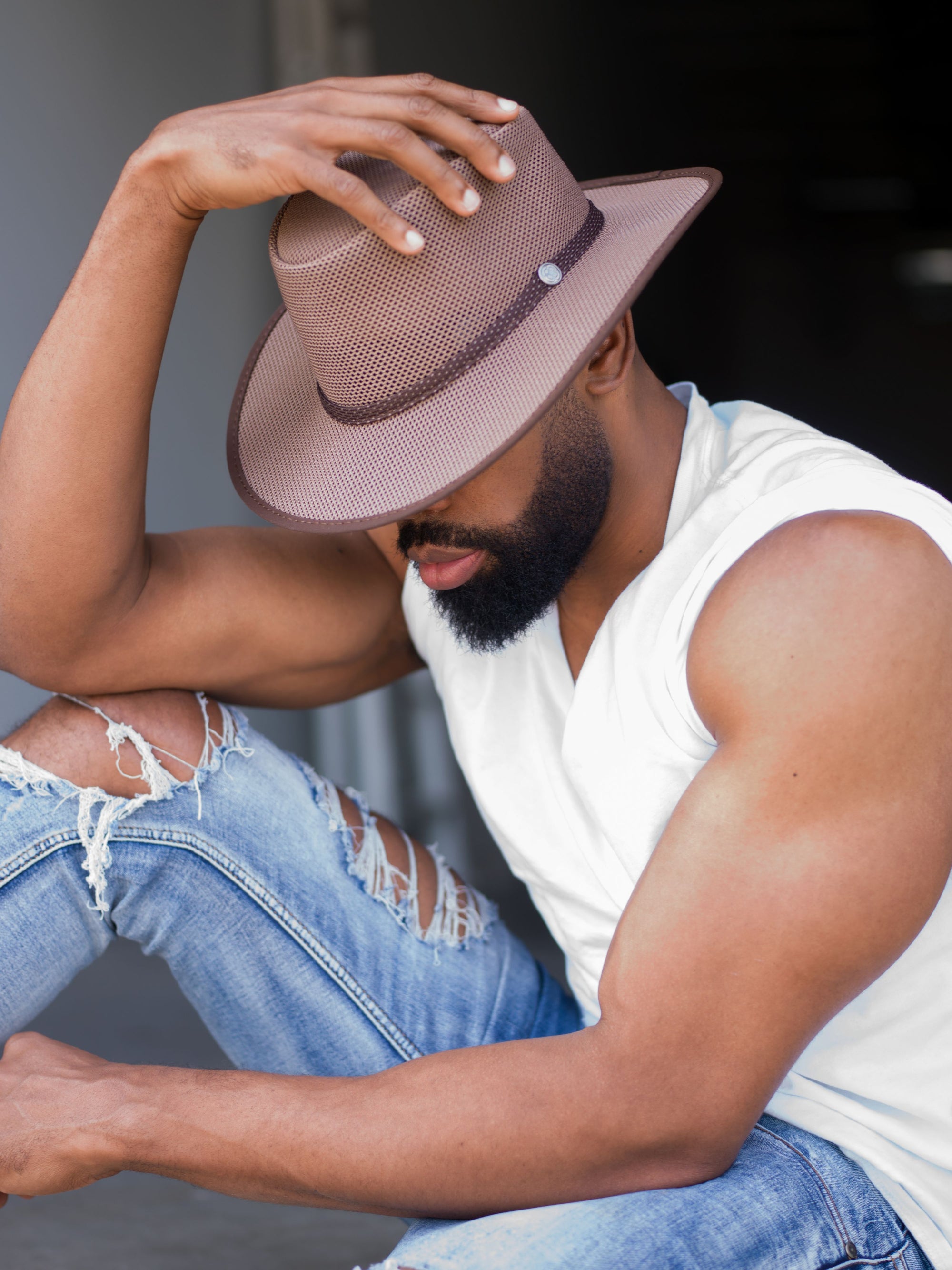 A man sitting down wearing tattered jeans and a brown sun hat