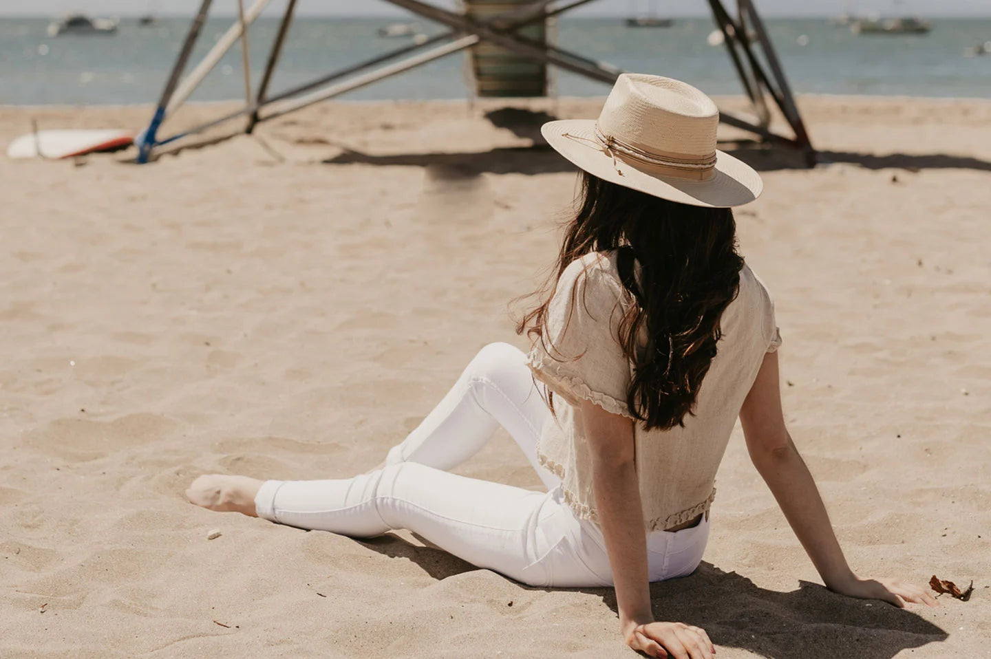 A woman sitting on the sand wearing a tan shirt, white jeans and a straw sun hat
