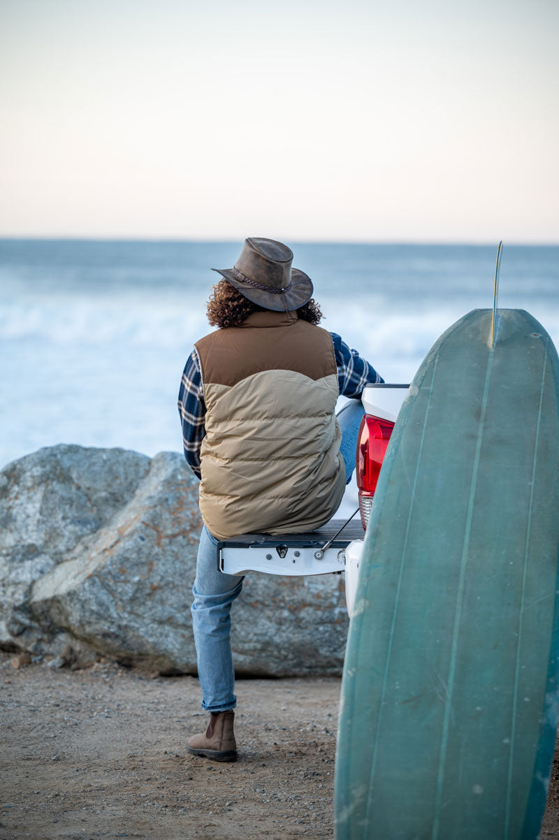 A man on the beach sitting on a truck wearing a bomber brown leather hat