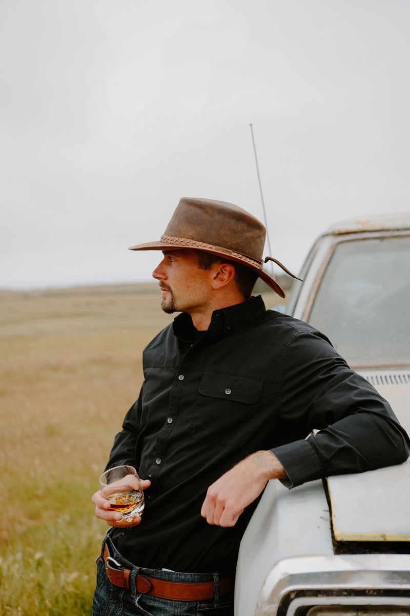 A man leaning on a truck while holding a glass and wearing a copper leather hat
