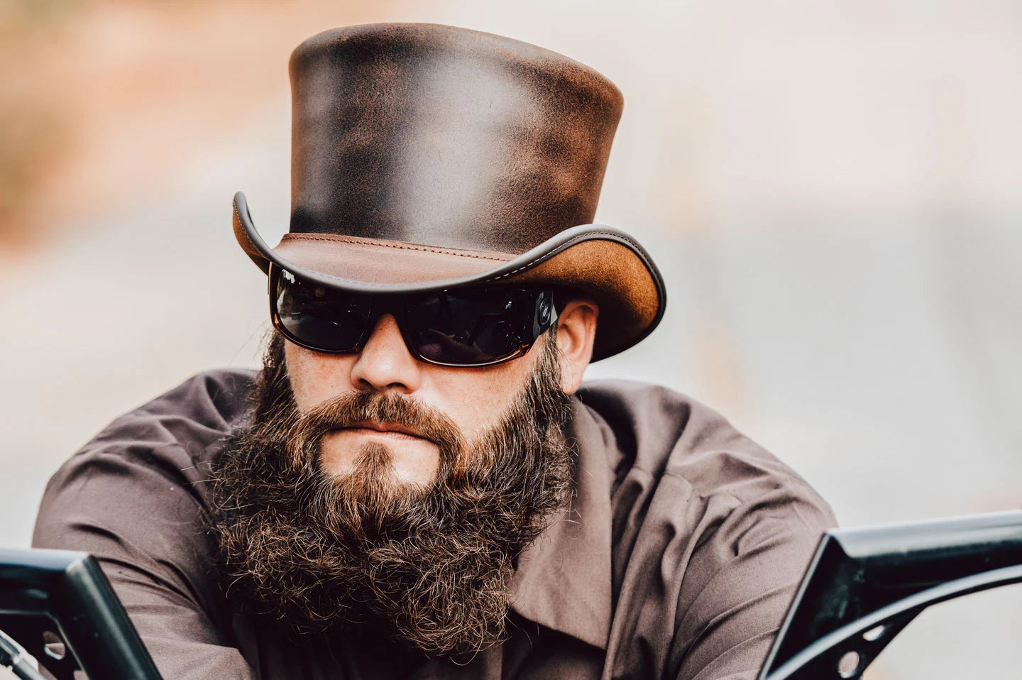 biker with a beard leaning on his harley davidson wearing a brown leather top hat
