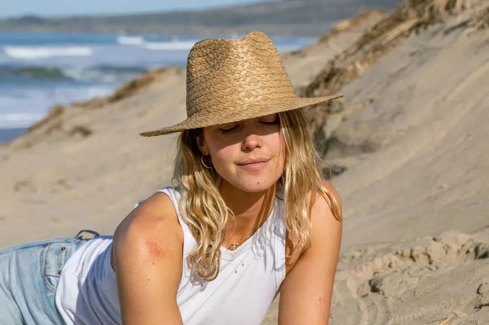 A woman relaxing on the beach wearing a white top, jeans and a straw sun hat