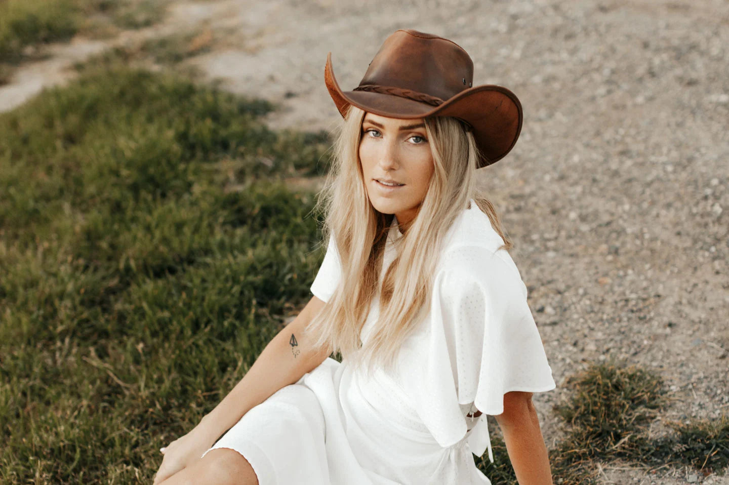A woman in a white dress sitting on the ground wearing a brown leather hat