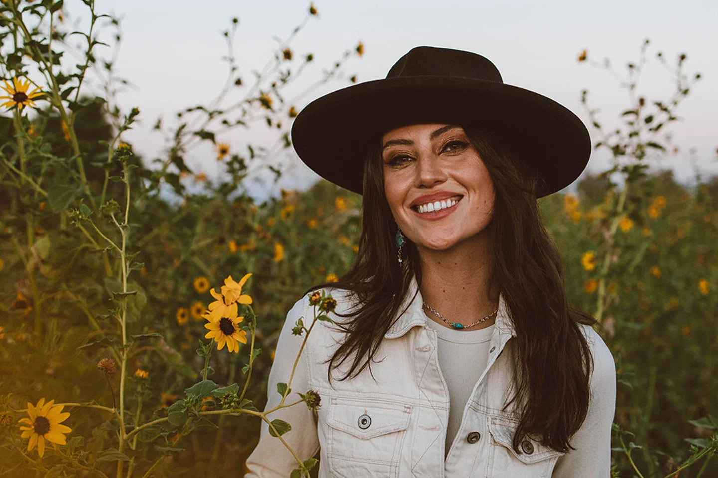 woman in a white jacket standing in a garden full of sunflowers wearing a black hat by american hat makers