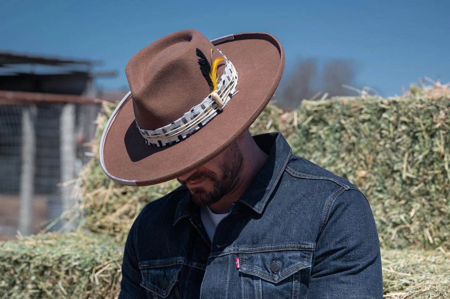 man wearing denim jacket next to hay bales wearing a brown felt fedora hat 