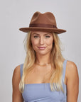 Portrait of a smiling woman with blonde hair, wearing a brown Pablo fedora and a blue sleeveless top.