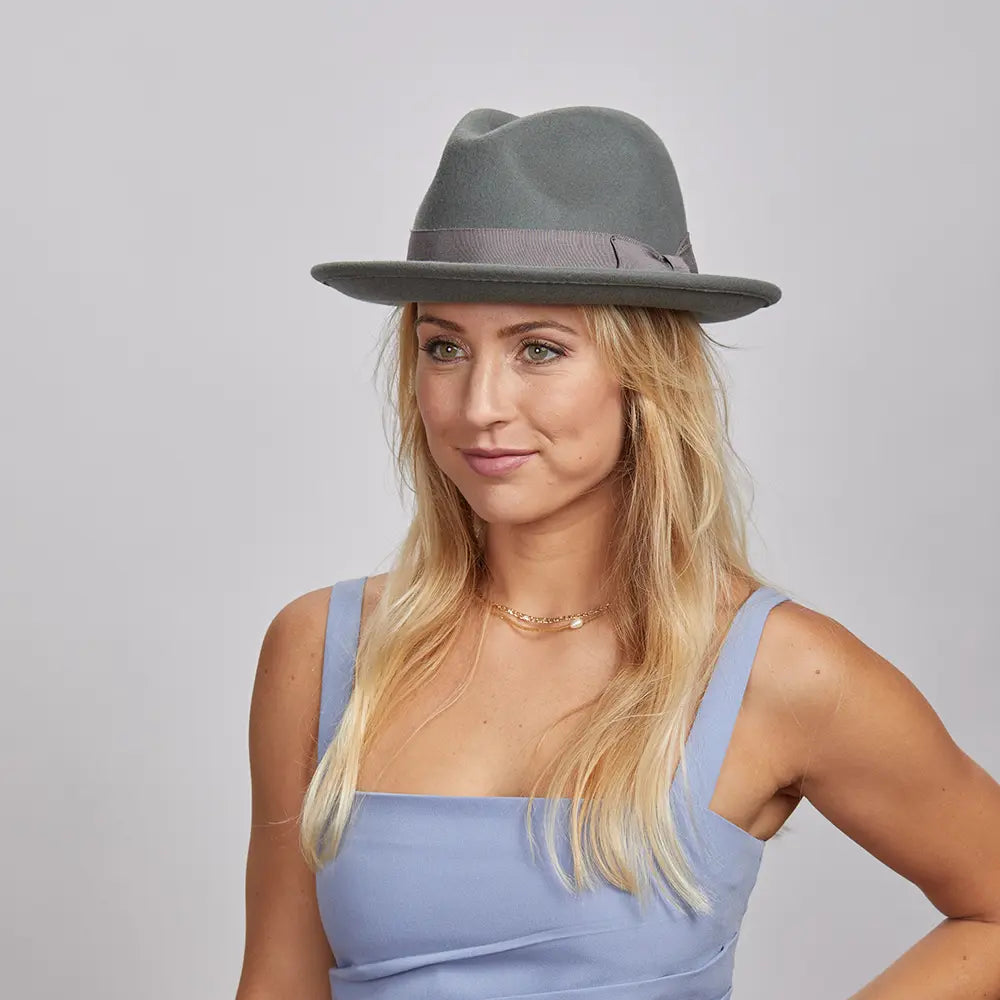 A smiling woman with blonde hair, wearing a grey Pablo fedora and a blue sleeveless top.