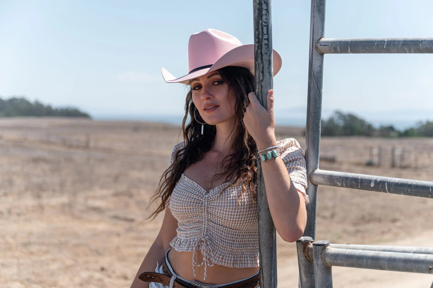 woman with a curly hair leaning in a metal fence wearing a pink hat by american hat makers