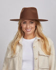 A woman with long blonde hair, smiling and wearing a brown Rancher hat and a textured cream coat over a white shirt