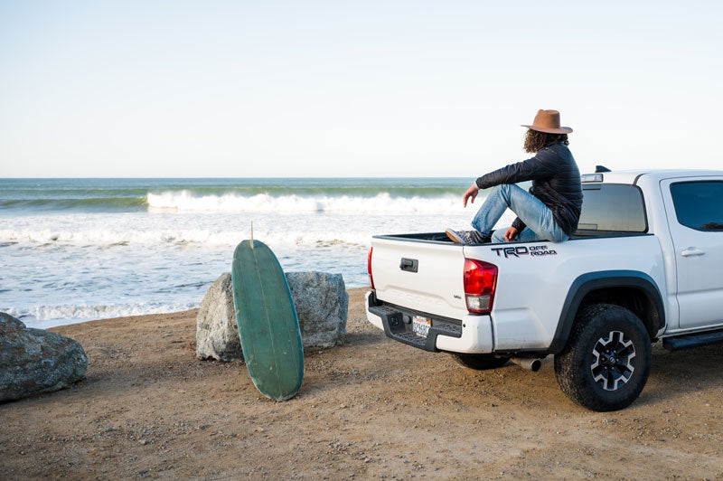 A man on the beach sitting on a truck wearing a brown felt fedora hat