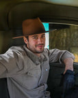 A man sitting inside a truck wearing a gray polo, gloves and brown felt hat