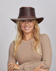 A smiling woman with blonde hair wearing a Chocolate Roughneck Outback Hat and a taupe long-sleeve top, standing with her arms crossed