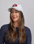 A woman with long brown hair smiling, wearing a white Stay Humble cap and a navy blue top.
