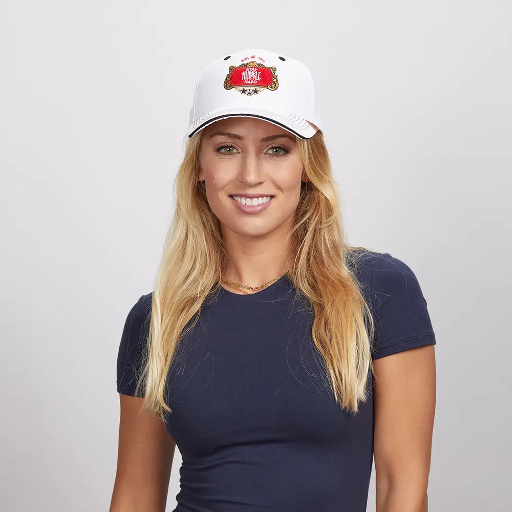 A blonde woman smiling, wearing a white Stay Humble cap and a navy blue top.