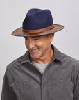 A man with a light stubble wearing a felt navy fedora hat and a gray button-up shirt.