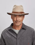 A man with a light stubble wearing an felt oatmeal fedora hat and a gray button-up shirt.