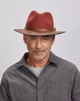 A man with a light stubble wearing a felt sangria fedora hat and a gray button-up shirt.