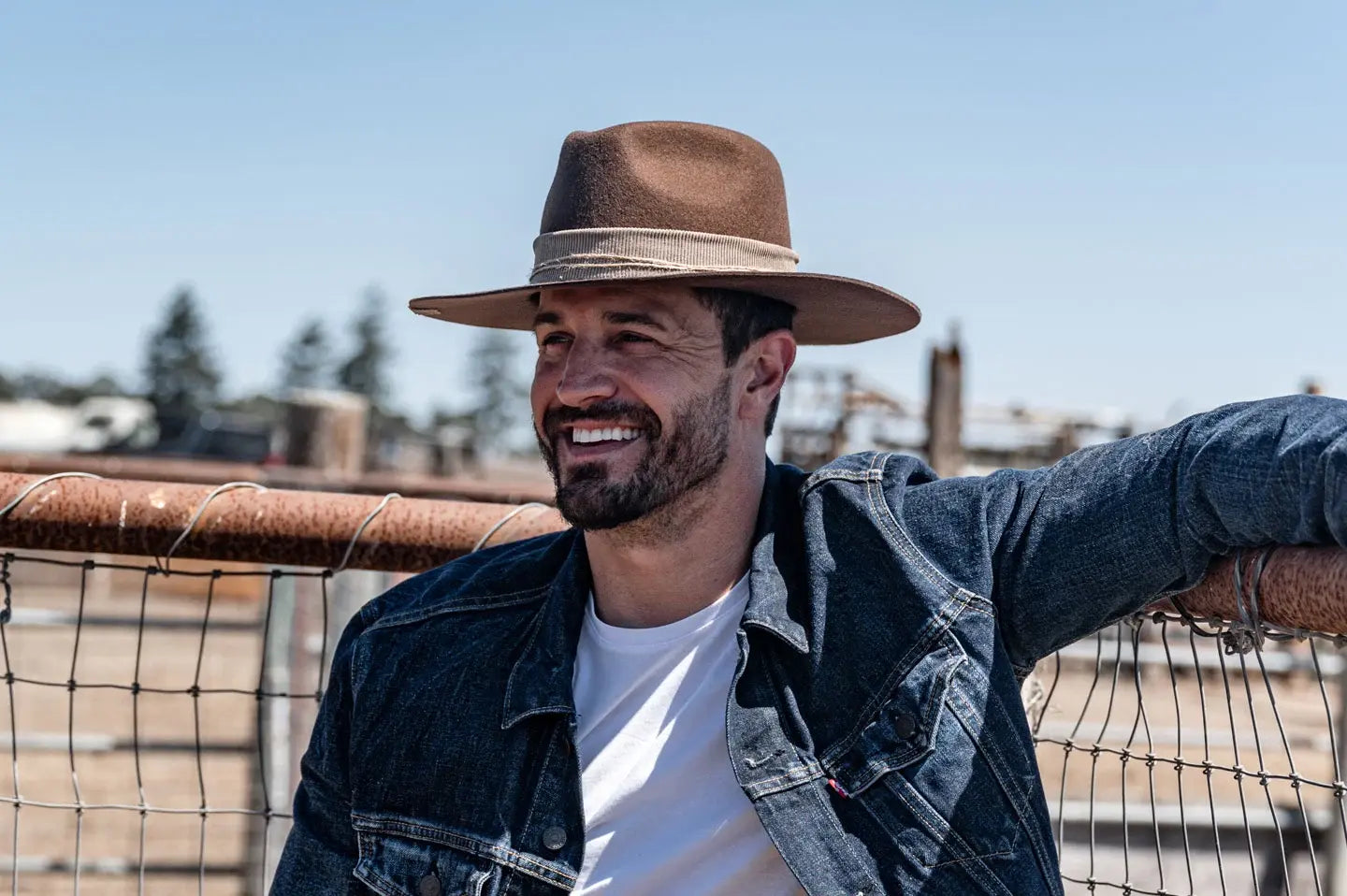 man leaning on a fence wearing a denim brown felt fedora hat 