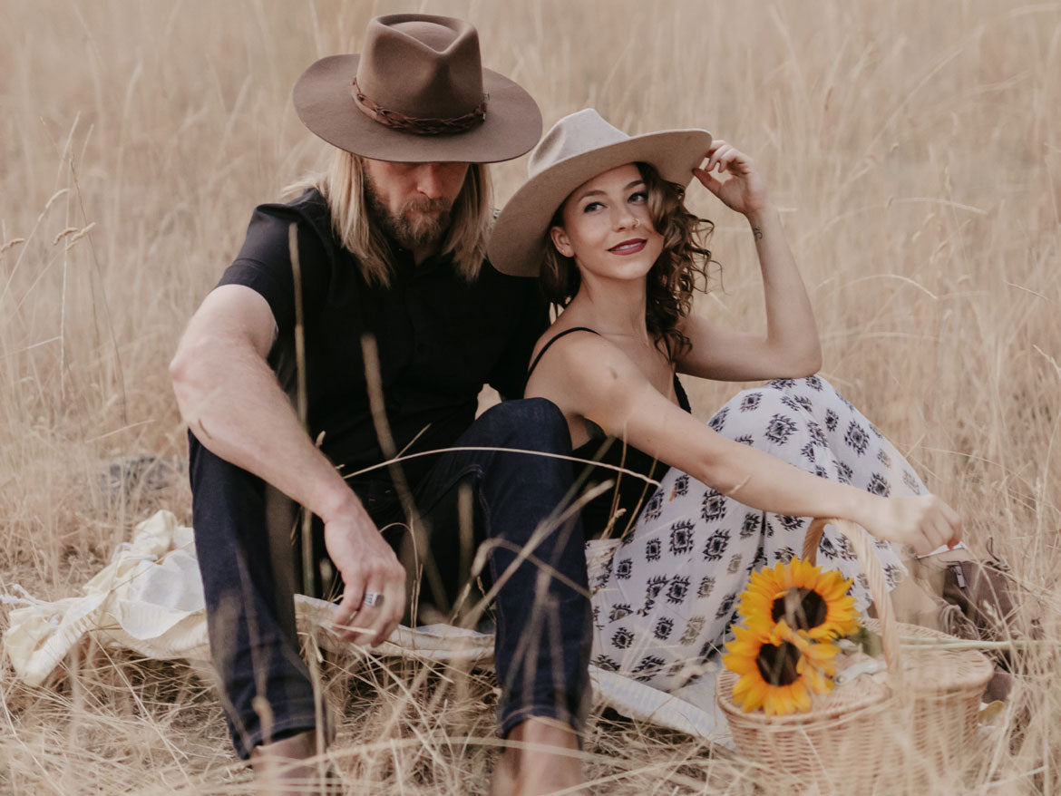 A couple having a picnic in an outdoor field wearing felt hats