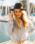 A woman standing on a boat fixing her hair wearing brown belt and a black hat