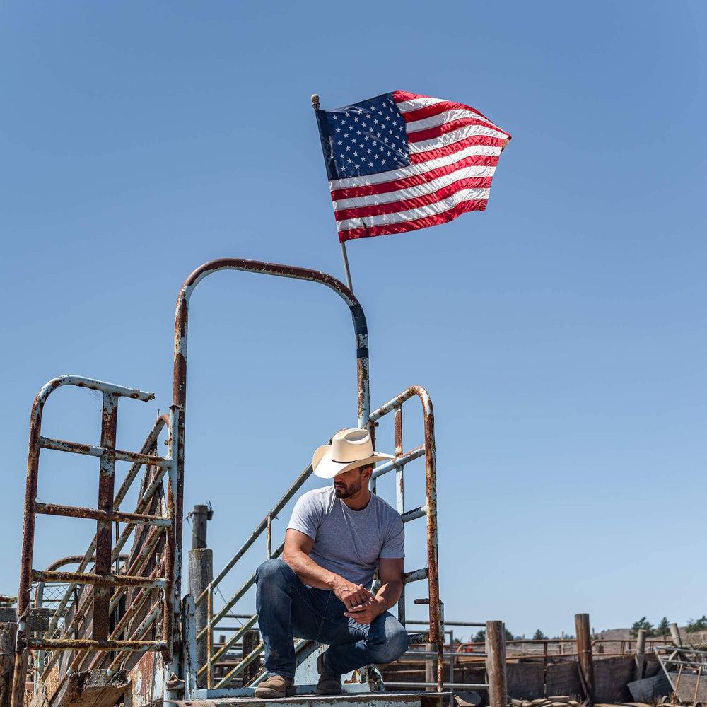A man squatting on a metal stairway with US flag above him and wearing Cream Straw Cowboy Hat