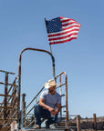 A man squatting on a metal stairway with US flag above him and wearing Cream Straw Cowboy Hat 