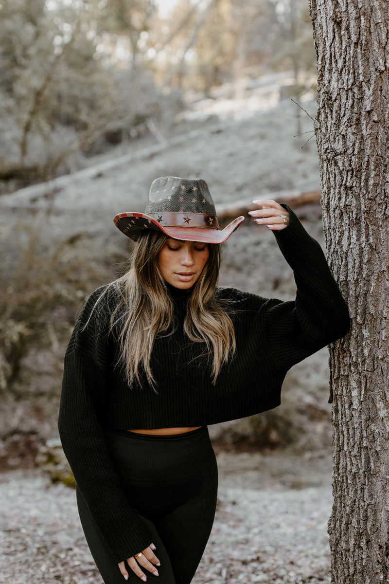 A woman leaning on a tree wearing a black outfit and a straw cowboy hat