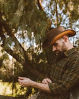 A man staying near a lake wearing a plaid shirt and brown leather hat