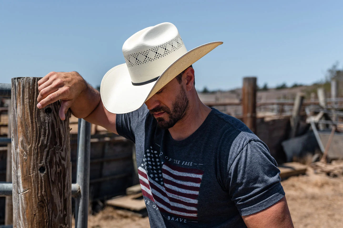 cowboy at a ranch leaning on a post wearing an american flag shirt wearing a straw cowboy hat
