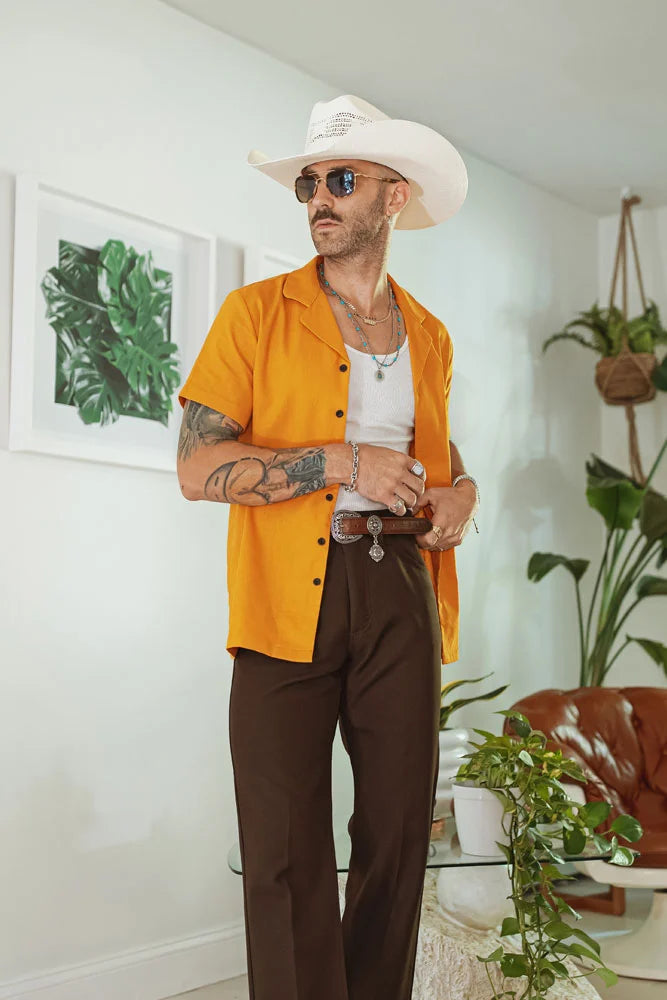 A man standing in a living room wearing an orange polo, white sleeveless, brown trousers and a cream cowboy hat