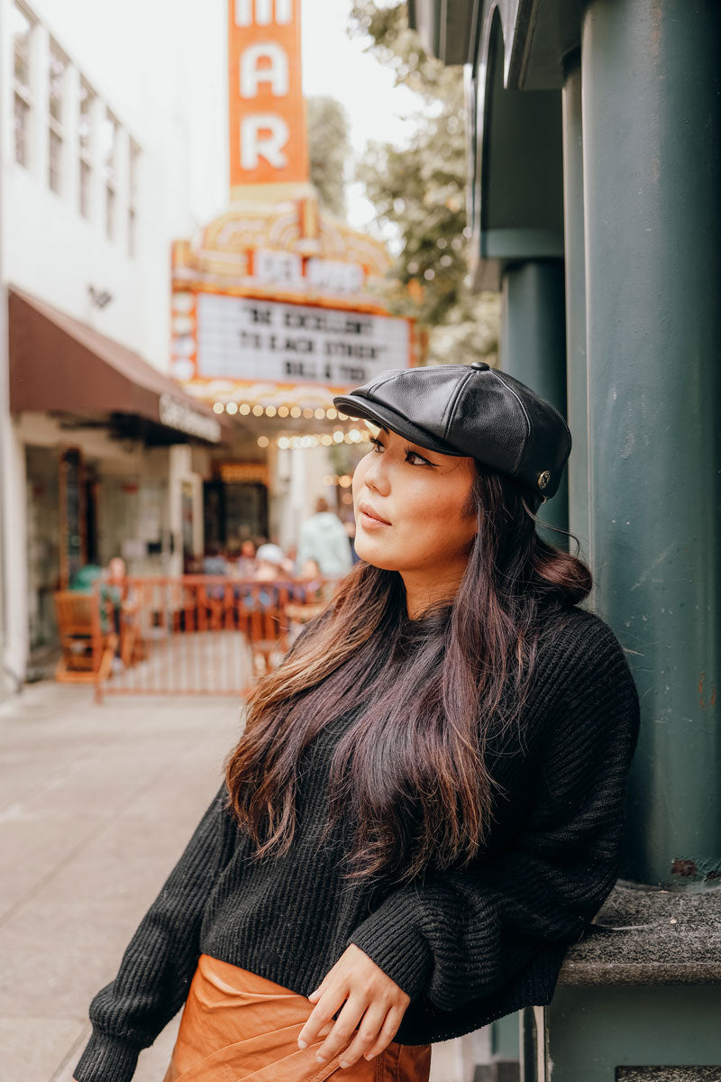 A woman on the  street leaning on the post wearing a black leather cap