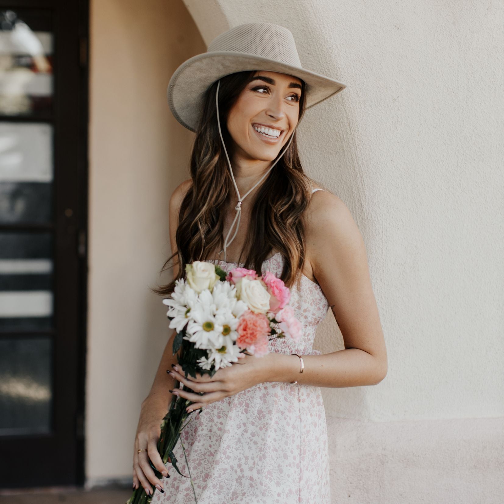 Woman holding flowers against wall while wearing the Breeze Latte sun hat