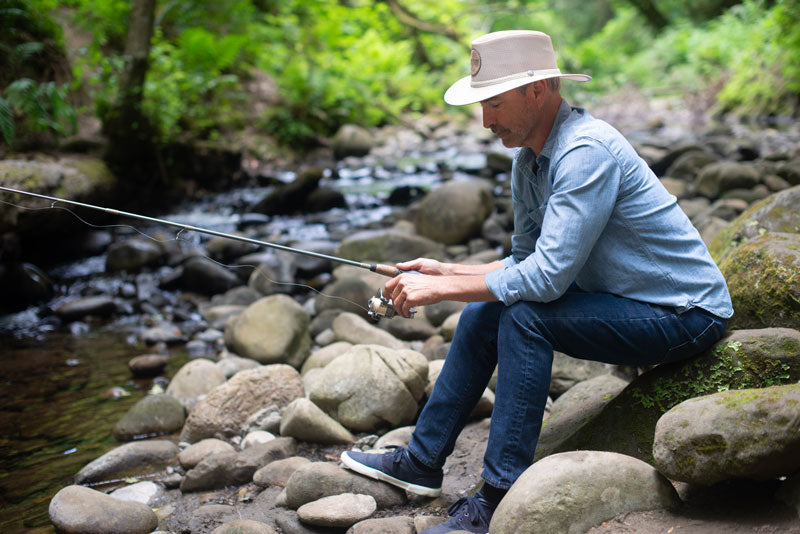 A man sitting down on a rock while fishing wearing an ivory logoed sun hat