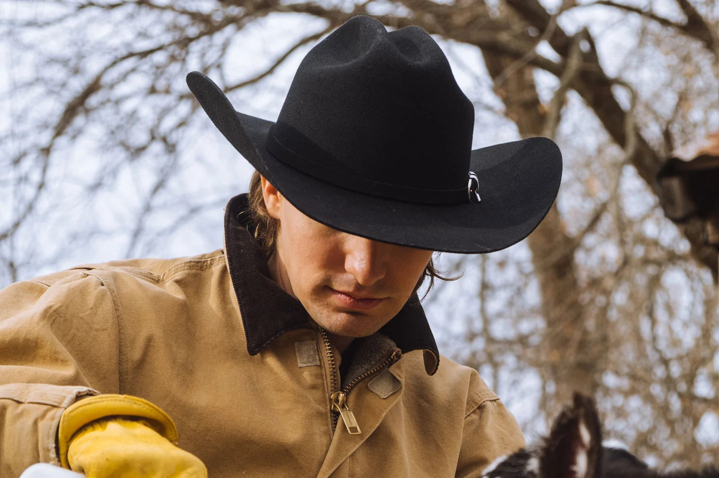 man in a brown jacket wearing gloves in the woods wearing a black felt cowboy hat by american hat makers