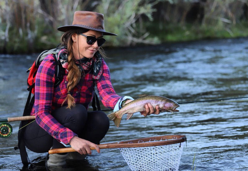 A woman in a checkered polo catching a fish and wearing a bomber brown leather hat