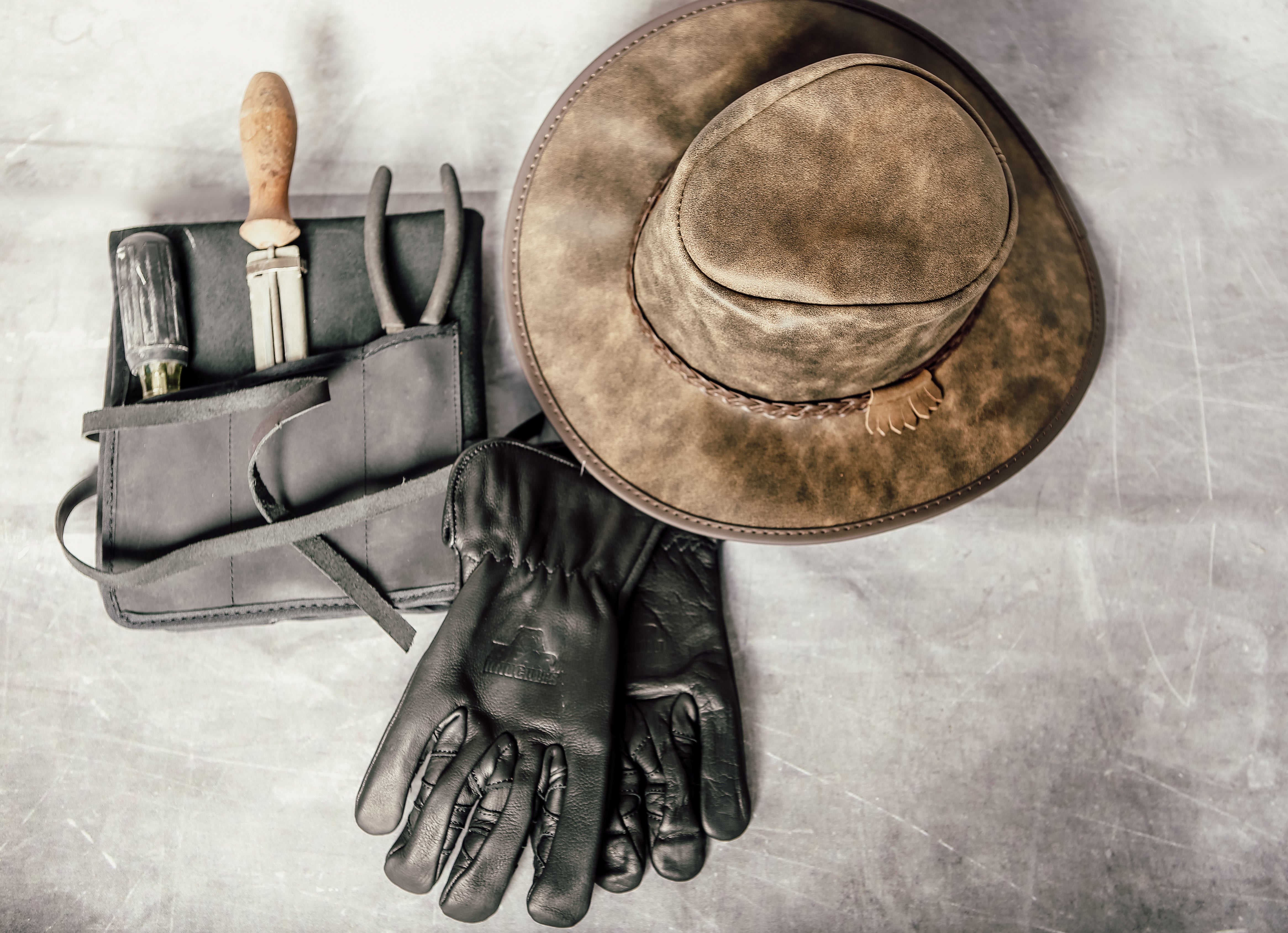 A brown cowboy hat together with 1 pair of black gloves and a tool wrap with tools