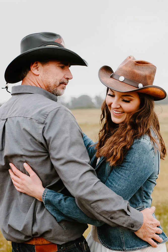 A man in a gray polo with black cowboy hat embracing a woman wearing a denim jacket and a brown leather cowboy hat