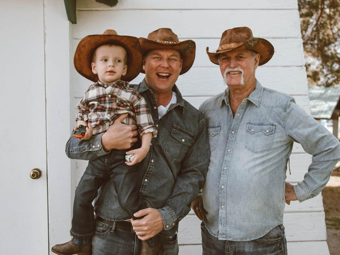 Two men with a child standing in front of a white house wearing brown leather cowboy hats