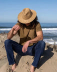 A man sitting on the shore wearing a brown t-shirt, denim jeans and a straw sun hat
