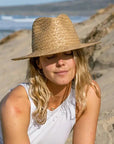 A woman relaxing on the beach wearing a white top, jeans and a straw sun hat 