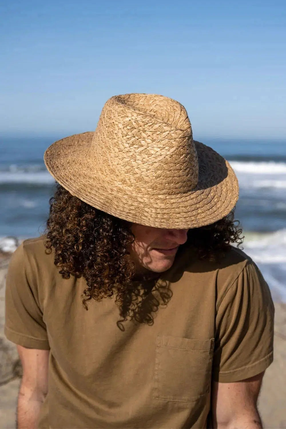 A curly haired man wearing a brown shirt and a straw sun hat sitting by the seashore