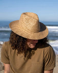 A curly haired man wearing a brown shirt and a straw sun hat sitting by the seashore