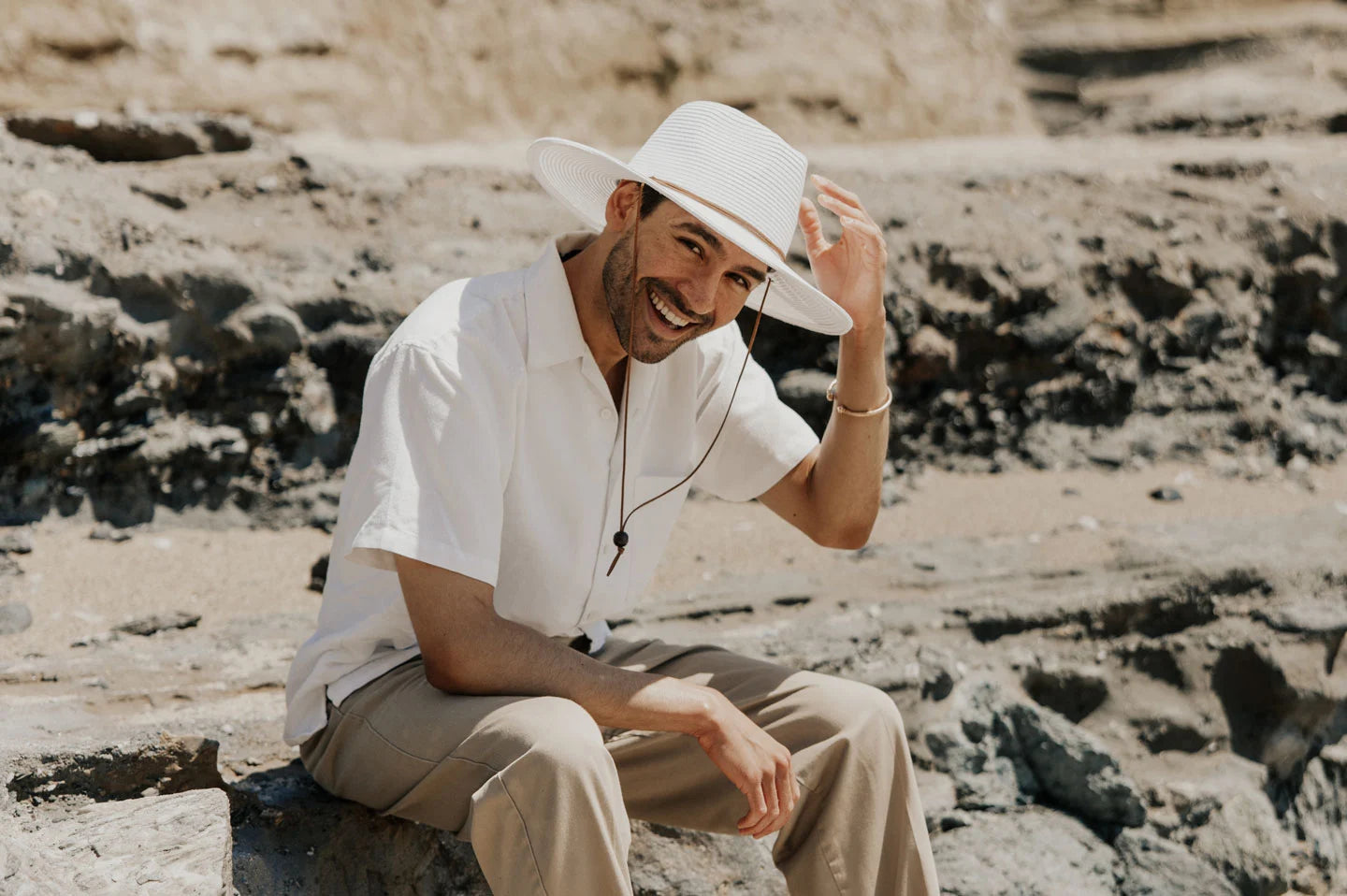 A man sitting on a rock wearing a polo shirt and a white sun hat with a chinstrap